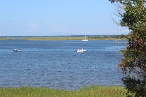 Two boats on a calm river with green marshland in the background and a sailboat in the distance under a clear blue sky.