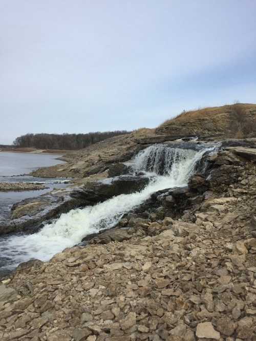 A waterfall cascades over rocky terrain, surrounded by a calm river and a distant tree line under a cloudy sky.