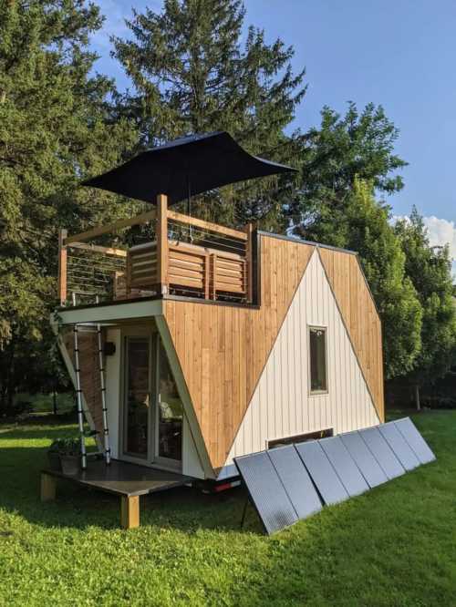 A modern tiny house on wheels with a wooden exterior, rooftop deck, and solar panels, set in a green landscape.