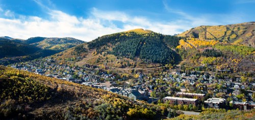 A panoramic view of a mountain town surrounded by vibrant autumn foliage and rolling hills under a clear blue sky.