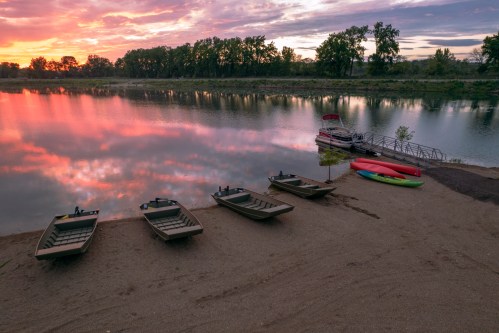 Boats lined up on a sandy shore at sunset, reflecting colorful skies on calm water.