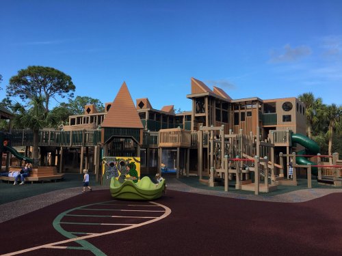 A large wooden playground with slides and climbing structures, surrounded by trees and a blue sky.