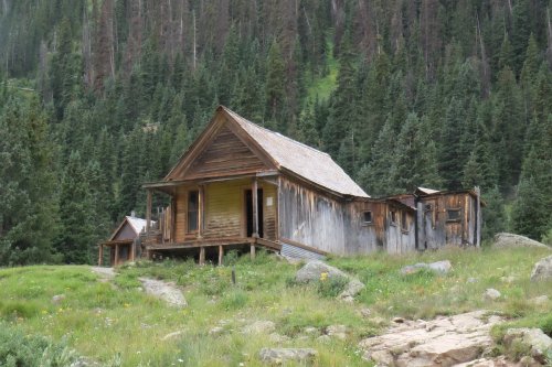 Abandoned wooden house surrounded by tall trees and grassy terrain in a mountainous area.