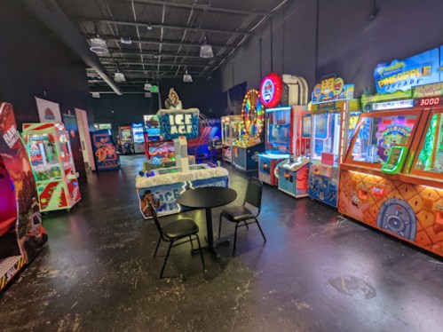 A vibrant arcade filled with various games and machines, featuring colorful lights and a central table with chairs.