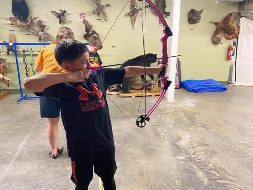 A young boy aims a pink bow and arrow in an indoor archery range, surrounded by mounted animal displays.