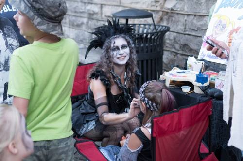 A performer in costume applies face paint to a child, surrounded by onlookers in a festive outdoor setting.