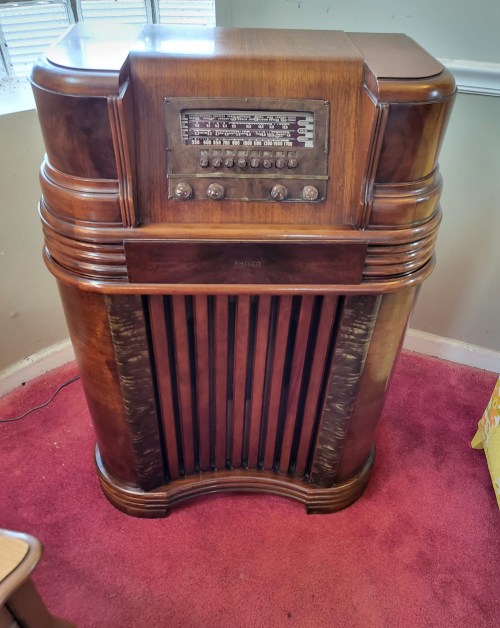 A vintage wooden radio console with a curved design and multiple knobs, set against a carpeted floor.