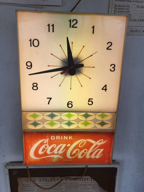 A vintage illuminated clock featuring a Coca-Cola advertisement at the bottom, with bold numbers and a classic design.
