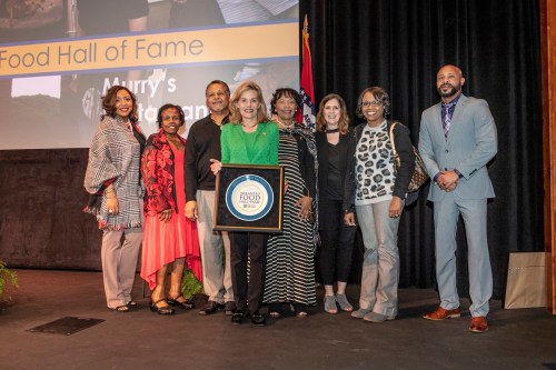 A group of eight people poses together on stage, holding a plaque for the Food Hall of Fame.
