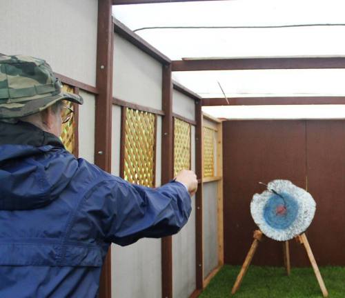 A person in a camo hat throws a dart at a target with a blue and white bullseye in a covered outdoor area.
