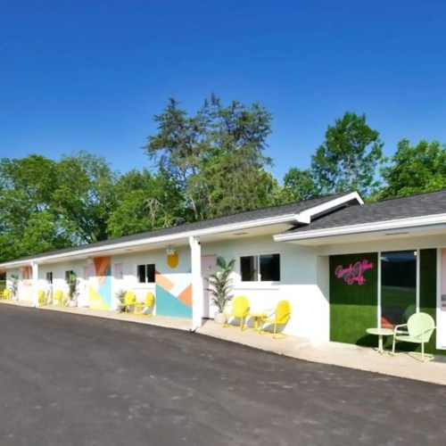 Colorful motel exterior with a curved driveway, green trees, and yellow chairs on the porch. Bright and inviting atmosphere.