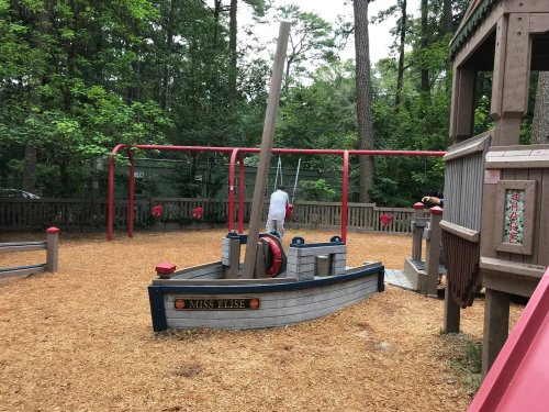 A playground featuring a wooden pirate ship, swings, and a slide, surrounded by trees and wood chips.
