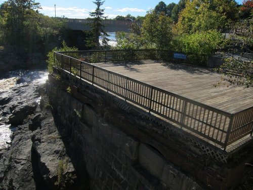 A wooden observation deck overlooking a river, surrounded by trees and rocky terrain under a clear blue sky.
