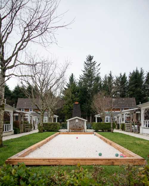 A bocce ball court surrounded by trees and cottages, with colorful balls scattered on the gravel surface.