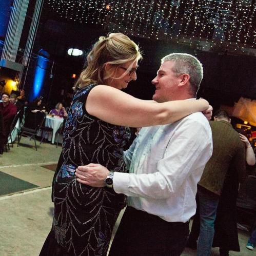 A couple dances closely together at a festive event, surrounded by twinkling lights and a joyful atmosphere.