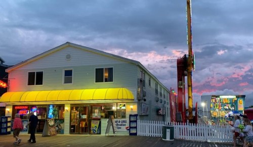 A colorful boardwalk scene at dusk, featuring a restaurant with a yellow awning and a nearby amusement ride.