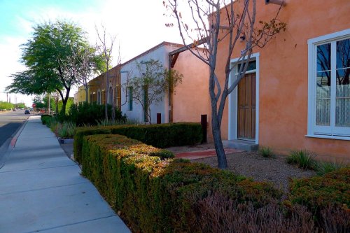 A sidewalk lined with neatly trimmed hedges and colorful adobe-style buildings under a clear blue sky.