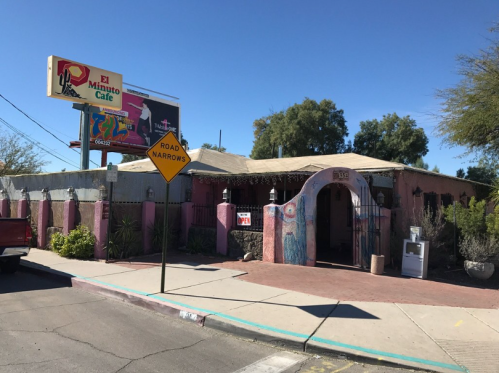 Colorful exterior of El Minuto Cafe with a sign, surrounded by desert vegetation and a "Road Narrows" warning sign.