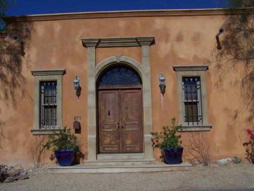 A rustic adobe-style building with a wooden double door, arched window, and blue planters on either side.
