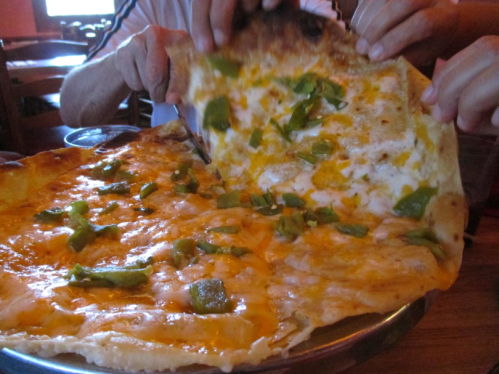 Two hands pulling apart a large pizza topped with melted cheese and green peppers on a metal tray.