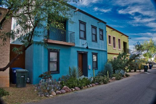 Colorful houses line a street, featuring a blue building with a balcony and a green building, surrounded by desert plants.