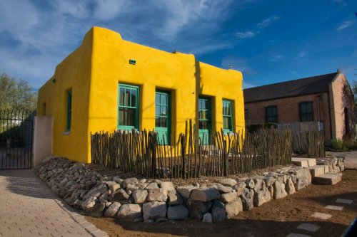 Bright yellow adobe house with turquoise windows, surrounded by a rock garden and cacti under a blue sky.