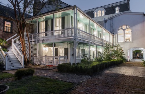 A charming two-story house with a wraparound porch, surrounded by greenery and illuminated at dusk.