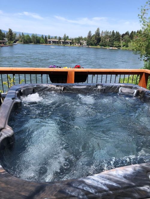 A hot tub overlooking a serene lake, surrounded by trees and a clear blue sky.