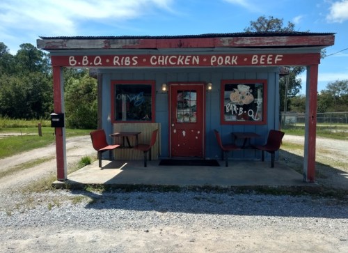 A small, rustic BBQ restaurant with a red and blue exterior, featuring outdoor seating and a sign that reads "BBQ."