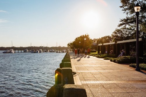 A sunny waterfront scene with people walking along a paved path beside a river and boats in the background.