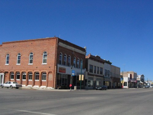 A quiet street scene featuring brick buildings and shops under a clear blue sky. A vintage car is parked nearby.