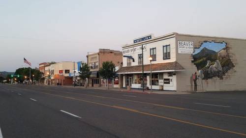 A quiet street scene featuring small shops and a mural, with trees and an American flag in the background.
