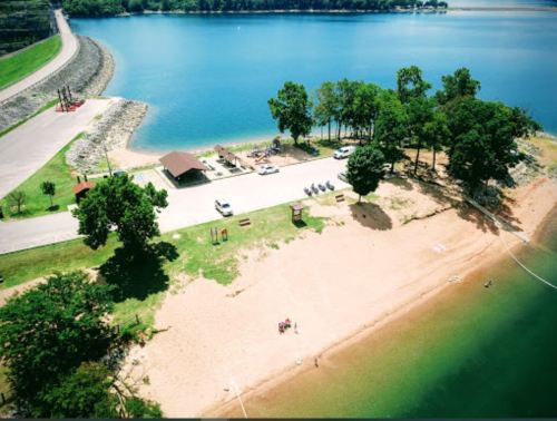 Aerial view of a sandy beach by a lake, with trees, picnic area, and parked cars along the shore.