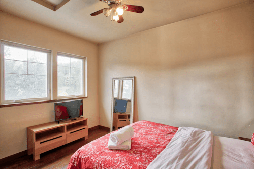 A cozy bedroom featuring a red bedspread, a mirror, a TV on a wooden stand, and two windows letting in natural light.