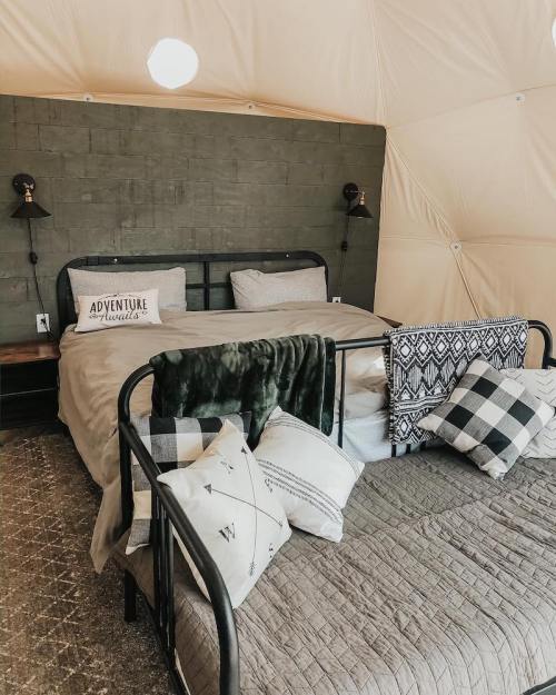 Cozy tent interior with two beds, decorative pillows, and a "Adventure Awaits" sign on the wall. Warm, inviting atmosphere.