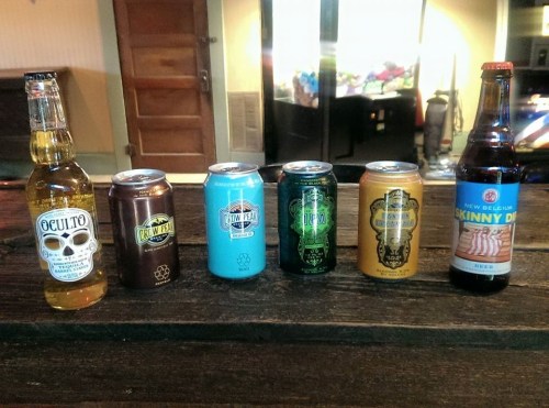 A lineup of various beer cans and bottles on a wooden surface, with a blurred background of a room.