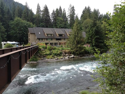 A rustic building by a river, surrounded by trees and greenery, with a bridge in the foreground.