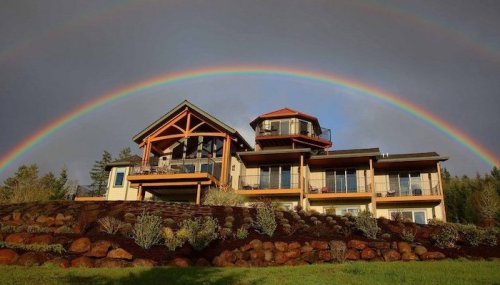 A modern house on a hillside with a rainbow arching over it, surrounded by greenery and rocky landscaping.
