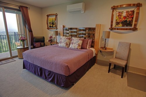 Cozy bedroom with a purple bedspread, decorative pillows, and artwork, featuring a balcony view and stylish furnishings.