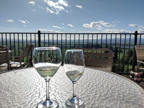 Two glasses of white wine on a table with a scenic view of green fields and mountains under a blue sky.