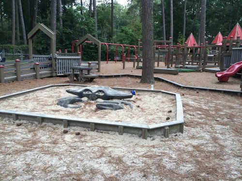 A playground with wooden structures, a sandy area featuring a rock formation, and trees in the background.