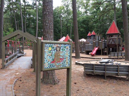 A playground with red-roofed structures, slides, and a mosaic sign surrounded by tall trees and pine needles.