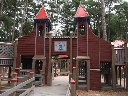 A colorful wooden playground structure with towers, surrounded by trees and a sandy area.