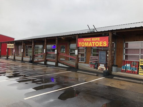 A rustic building with a sign reading "Vine Ripe Tomatoes," featuring a covered porch and wet pavement.