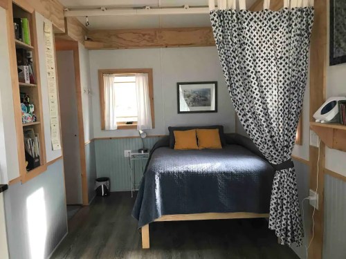 Cozy interior of a small room featuring a bed, curtains, and shelves with books and decor.
