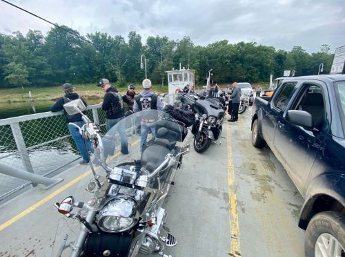 A group of motorcyclists on a ferry, with bikes parked and a river in the background, surrounded by trees.