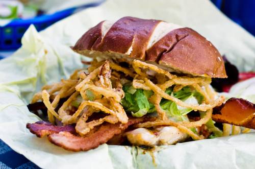 A close-up of a pretzel bun sandwich filled with lettuce, crispy onions, and bacon, served on green paper.