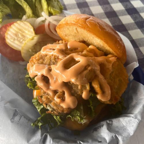 A close-up of a fried chicken sandwich with lettuce and sauce, served with pickles, onions, and tomato on the side.