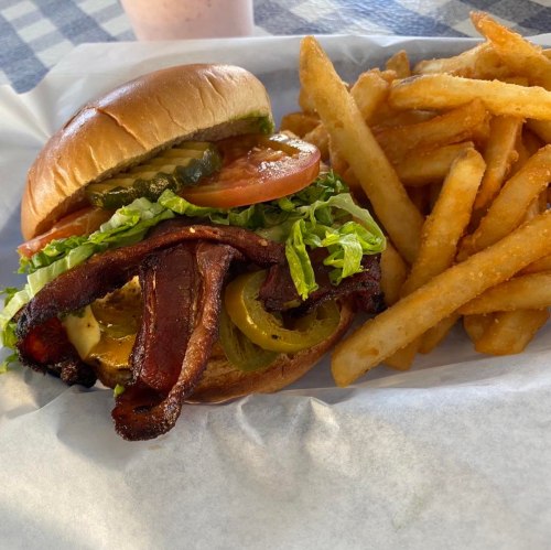 A juicy burger with bacon, lettuce, tomato, pickles, and fries on the side, served on a paper-lined plate.