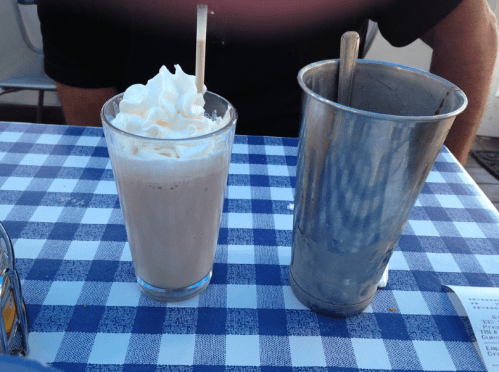 A glass of milkshake topped with whipped cream next to a metal shaker on a checkered tablecloth.